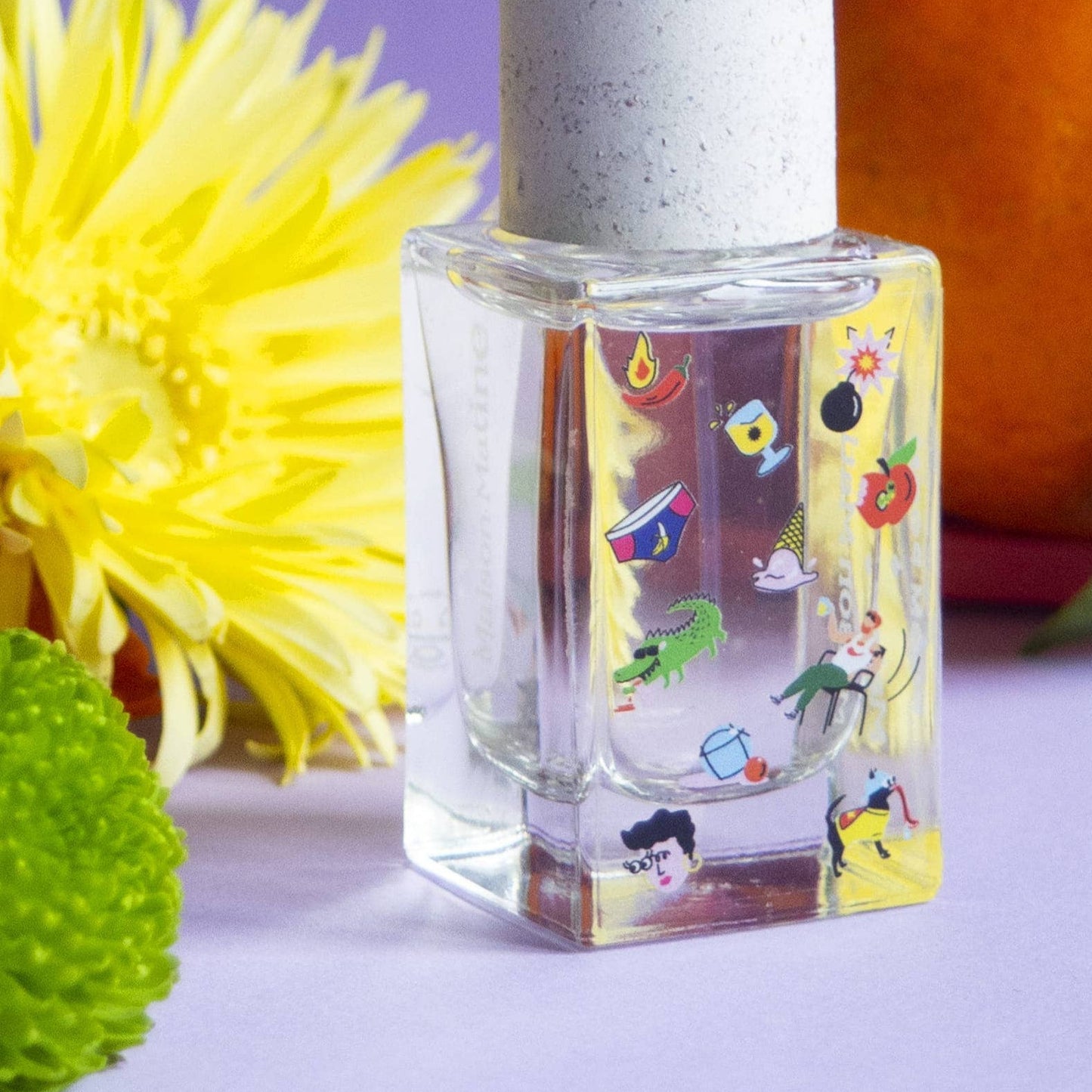 A clear glass bottle of perfume with a colorful label, accompanied by a red basket and a decorative flower, set against a purple background.