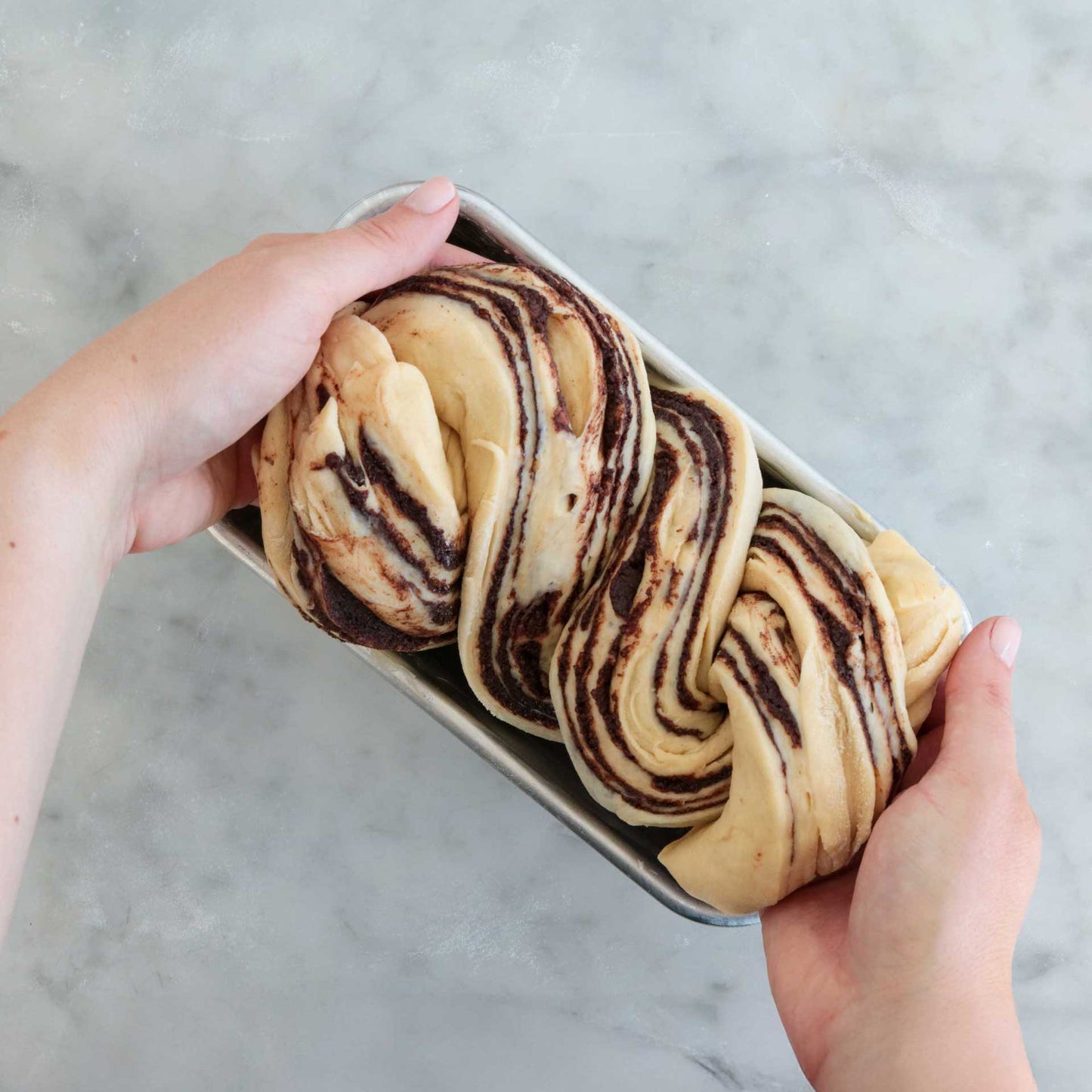 Braided bread with chocolate swirls in a loaf pan held by two hands on a marble surface.